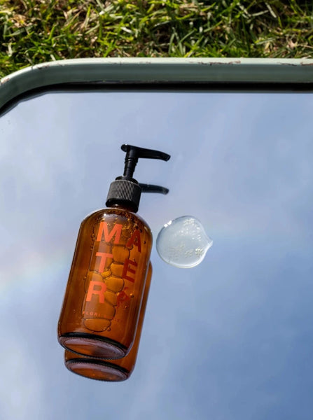 a glass bottle of flori liquid soap from mater lays next to some spilled soap on top of a mirror that reflects the blue sky
