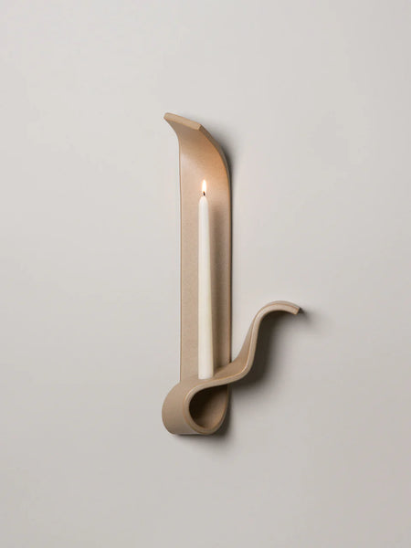 a tan ceramic wall sconce resembling a ribbon made by sin ceramics holding a lit taper  on ta white wall