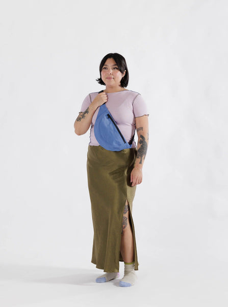 a woman carrying a pansy blue fanny pack over her shoulder with a black front zipper and black strap made by baggu against a white background 