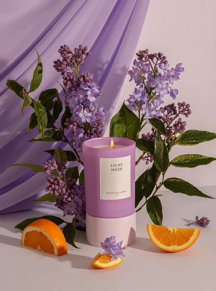 a frosted purple glass candle in the scent lilac haze by Brooklyn candle company sits next to pieces of orange and lilac