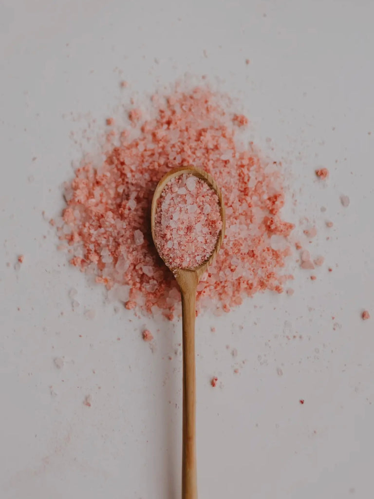 a wooden spoon full of bath salts sits on top of a pile of the same pink salt