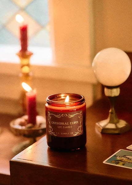 Candle in a glass jar with a label on a wooden surface, surrounded by other candles and decor.