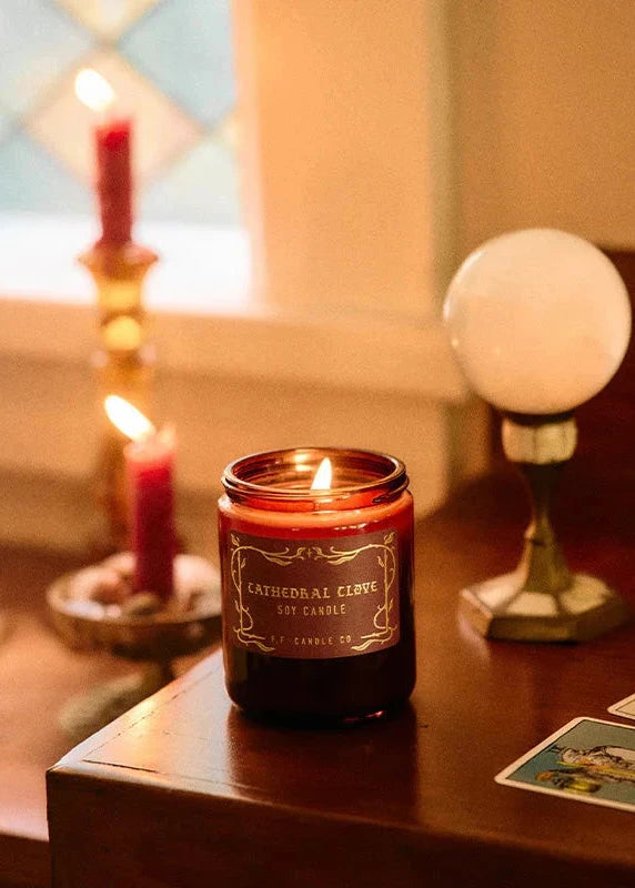 Candle in a glass jar with a label on a wooden surface, surrounded by other candles and decor.