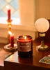 Candle in a glass jar with a label on a wooden surface, surrounded by other candles and decor.