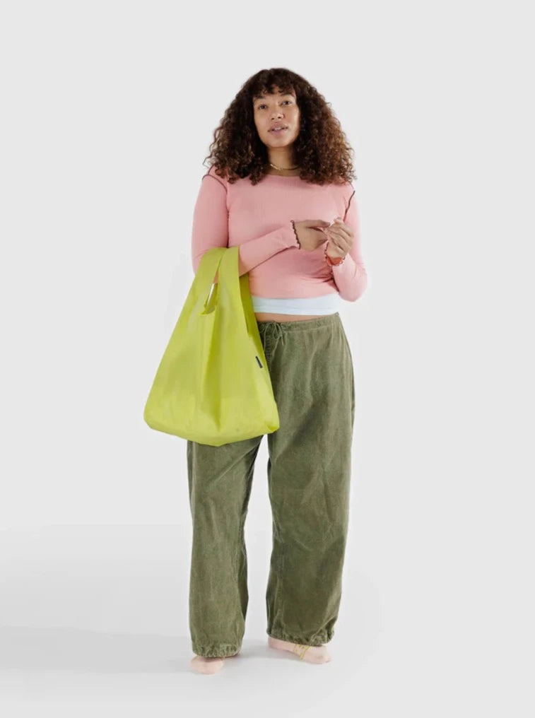 a woman in a pink shirt and green pants stands holding a lemon yellow reusable baggu bag on a white background