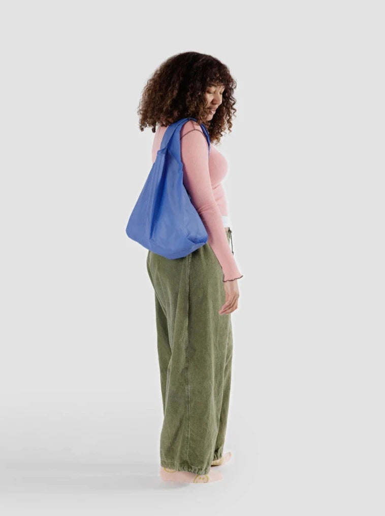 a woman in a pink shirt and green pants stands holding a cornflower blue reusable baggu bag on a white background