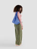 a woman in a pink shirt and green pants stands holding a cornflower blue reusable baggu bag on a white background