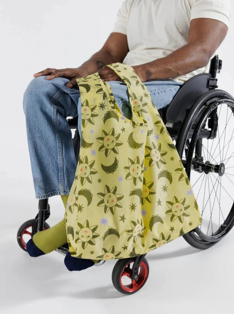 a man in a white shirt and blue jeans sitting in a wheelchair holding a yellow with green stars and suns patterned reusable baggu bag on a white background