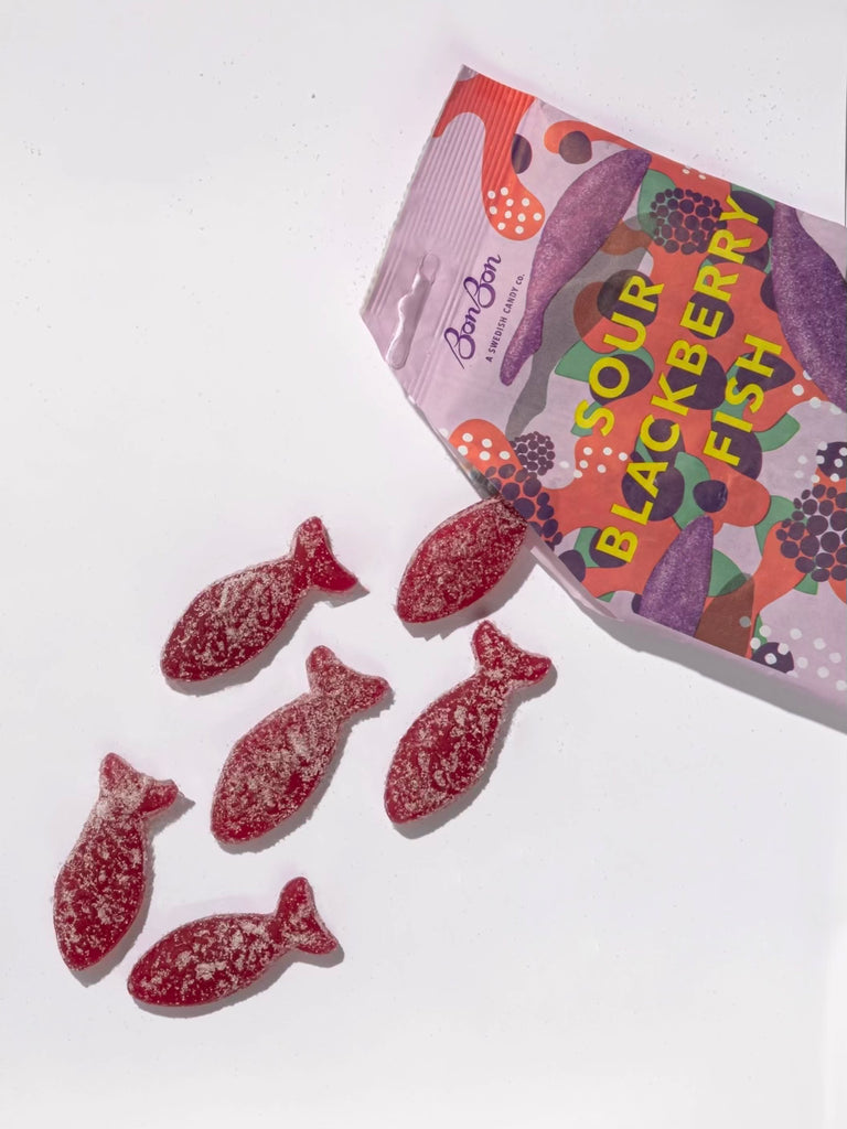 sour blackberry gummy fish spilling out of an opened illustrated bonbon nyc candy bag on a white background