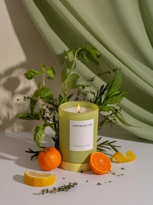 An Herbarium Candle from Brooklyn Candle Studio sits on a white tabletop, a sage curtain draped across the background. Around the candle are bundles of herbs such as basil, thyme and rosemary as well as sliced oranges, limes and citrus twists.