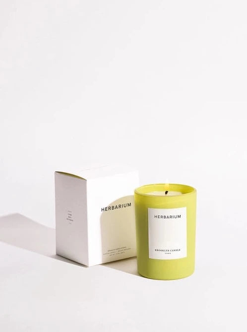 An Herbarium candle, in a bright chartreuse container, sits in front of a plain white background. A white box sits behind the candle.