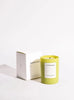 An Herbarium candle, in a bright chartreuse container, sits in front of a plain white background. A white box sits behind the candle.