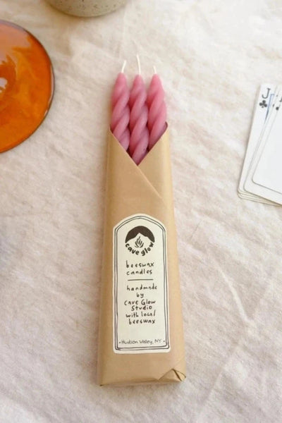 A set of three cherry colored spiral taper candles by Cave Glow Studio sits on a linen tablecloth. Around the candles are various items including a few playing cards, a yellow candle, a glass amber candle holder with a pale yellow spiral taper candle inside and a vase with white flowers inside. 