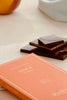 A broken dark chocolate peach bar from Cioccolato Lavoratti 1938 sits on a stone slab. The bar is broken to expose the peach filling. A closed box sits resting on the slab at an angle. 