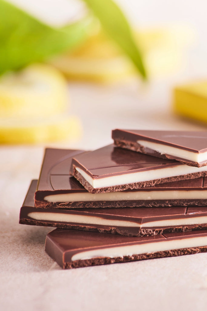 A stack of Cioccolato Lavoratti Lemon Cream filled Dark Chocolate Bars sit on top of a table. The bars are broken in half exposing the lemon cream filled center. There are a pile of lemons out of focus in the background. 