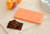 A broken dark chocolate peach bar from Cioccolato Lavoratti 1938 sits on a stone slab on top of a white table. The packaging for the chocolate bar sits next to it on the stone slab. Two peaches sit on the table above the chocolate on the stone slab. 