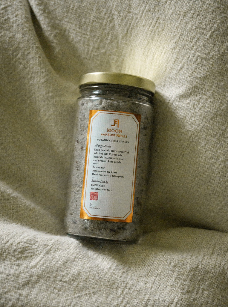 a glass jar of moon salts with rose petals sits on a cream blanket