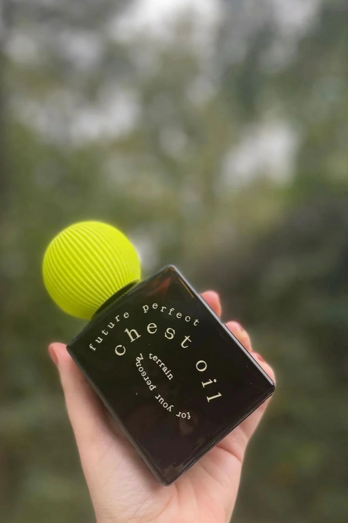 a hand holds a bottle of future perfect's chest oil in front of a background of hazy trees.