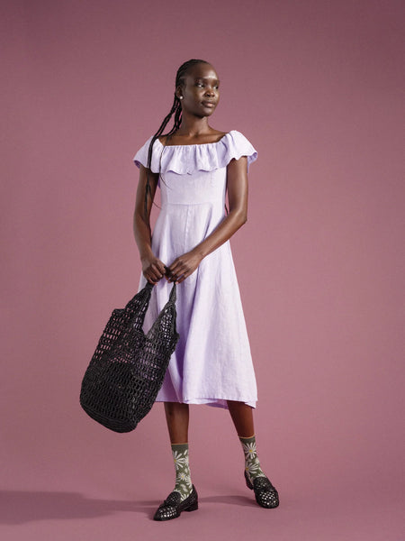 a woman in a lavender dress with colorful socks is standing in front of a pink background holding a black crocheted raffia bag made by Hansel from basel