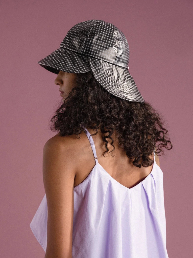 a woman with black curly hair is standing with her back to the camera in front of a pink background and wearing a lavender dress and a black and white gingham sun hat made by Hansel from Basel