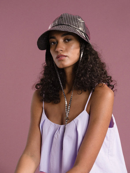 a woman with black curly hair is sitting in front of a pink background and wearing a lavender dress and a black and white gingham sun hat made by Hansel from Basel