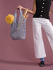 a woman in a black shirt and white pants with colorful socks is standing in front of a pink background holding a blue crocheted raffia bag with flowers and baguettes inside