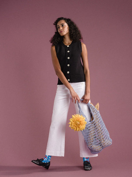 a woman in a black shirt and white pants with colorful socks is standing in front of a pink background holding a blue crocheted raffia bag with flowers and baguettes inside