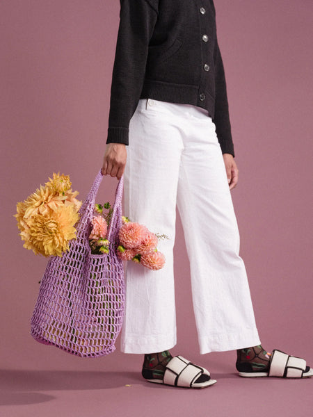a woman in a black shirt and white pants with colorful socks is standing in front of a pink background holding a lavender crocheted raffia bag with flowers and baguettes inside
