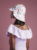 a woman standing with her back to the camera in front of a pink background and wearing a lavender dress and a white sun hat with colorful flowers made by Hansel from Basel