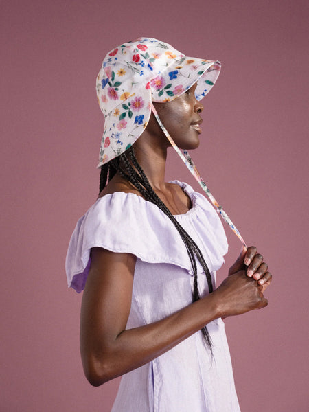 a profile of a woman standing in front of a pink background and wearing a lavender dress and a white sun hat with colorful flowers made by Hansel from Basel