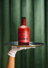 Red bottle with 'La Saum' branding on a black tray held by a gloved hand against a green curtain background.