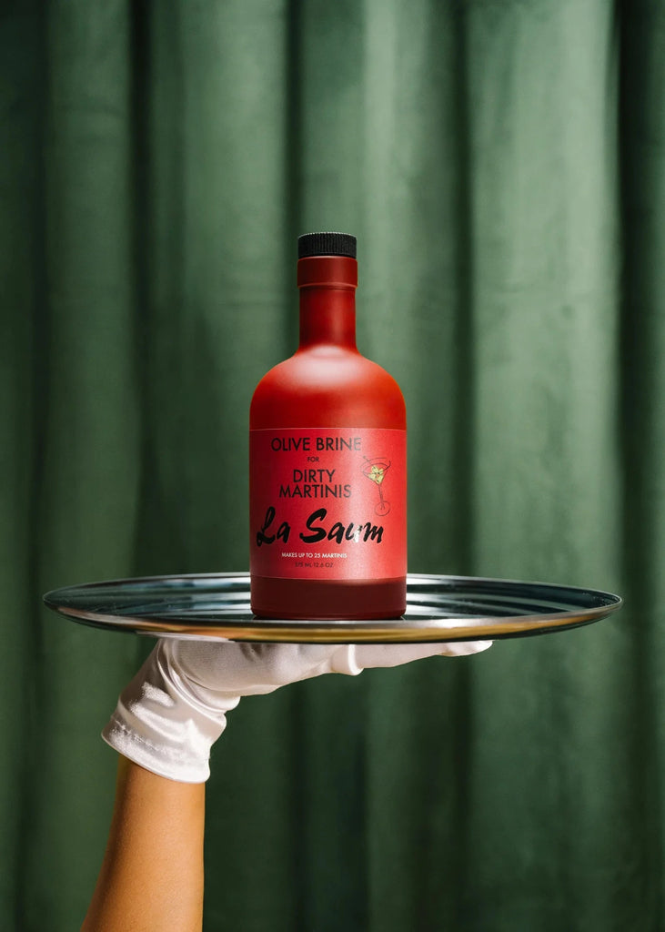 Red bottle with 'La Saum' branding on a black tray held by a gloved hand against a green curtain background.
