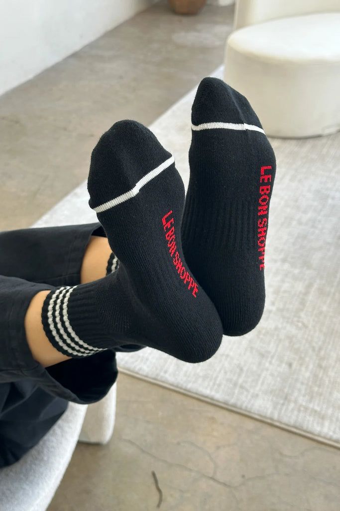 A woman lays with her back on a sofa, legs bent and holding her feet in the air. On her feet she wears a pair of black Girlfriend socks from Le Bon Shoppe