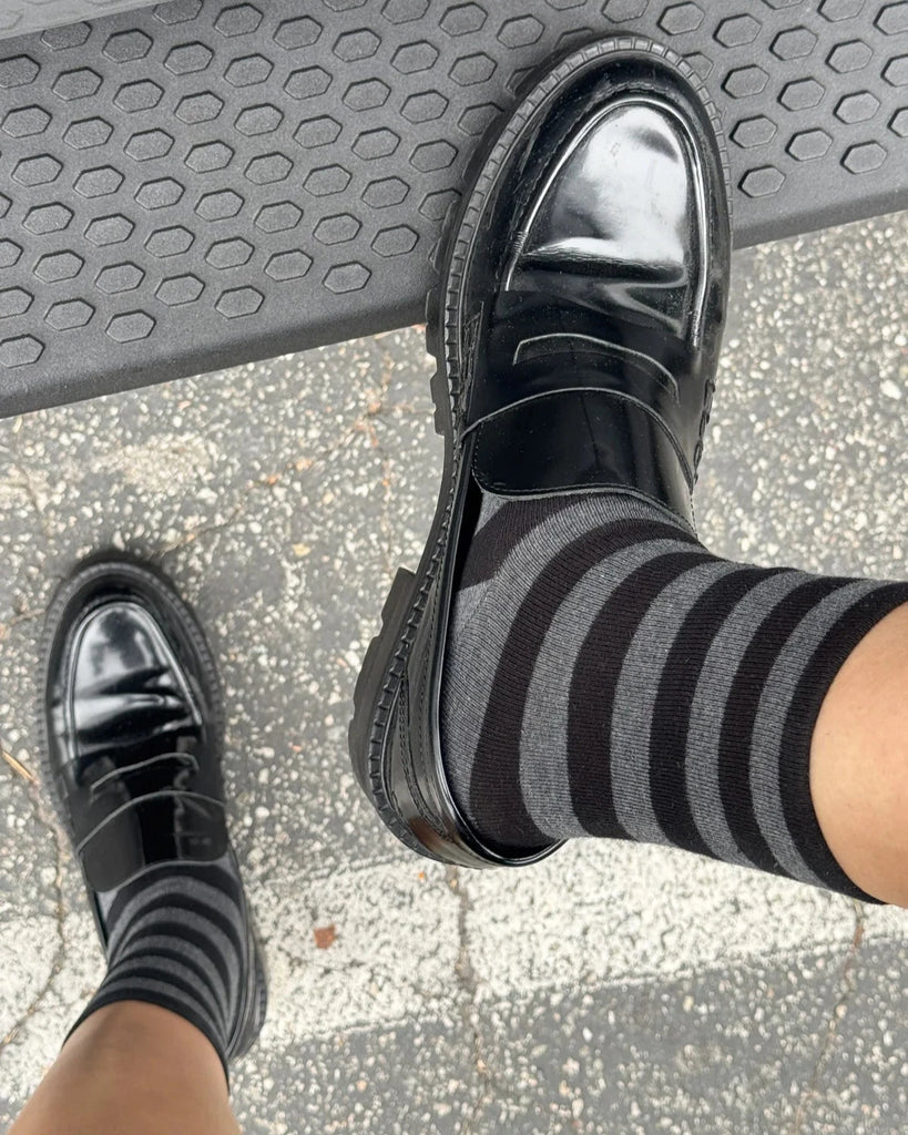 A person wearing a pair of Le Bon Shoppe Wally Socks in the color charcoal wide stripe paired with shiny loafers