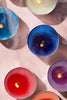 A close up shot of various different brightly colored tea light candles from Le Feu De L'eau sit on a stone table. The candles are lit, their smoke creating blurs across the photo. 