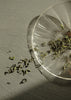 Dried green tea leaves on a clear glass plate with scattered herbs on a gray surface