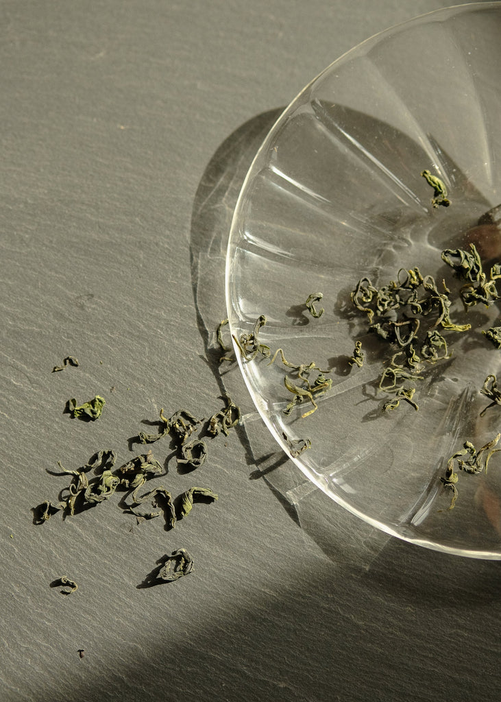 Dried green tea leaves on a clear glass plate with scattered herbs on a gray surface