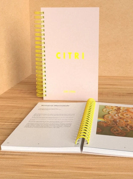 A Citri Cookbook sits standing straight up on a table in front of a light wooden background. An open Citri Cookbook sits on the table in front of the other book. A Kumquat Marmalade recipe is displayed on the left page with a photograph of sliced citrus paired on the other page.