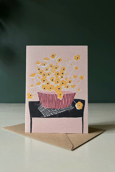 a pink greeting card with an illustration of yellow buttercups sitting on a black table by Luiza Holub.