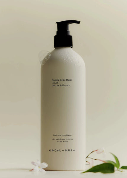 A pump bottle of Maison Louis Marie Bois de Balincourt body and hand wash with a label and branding visible, placed on a neutral background with some flowers and a green leaf.