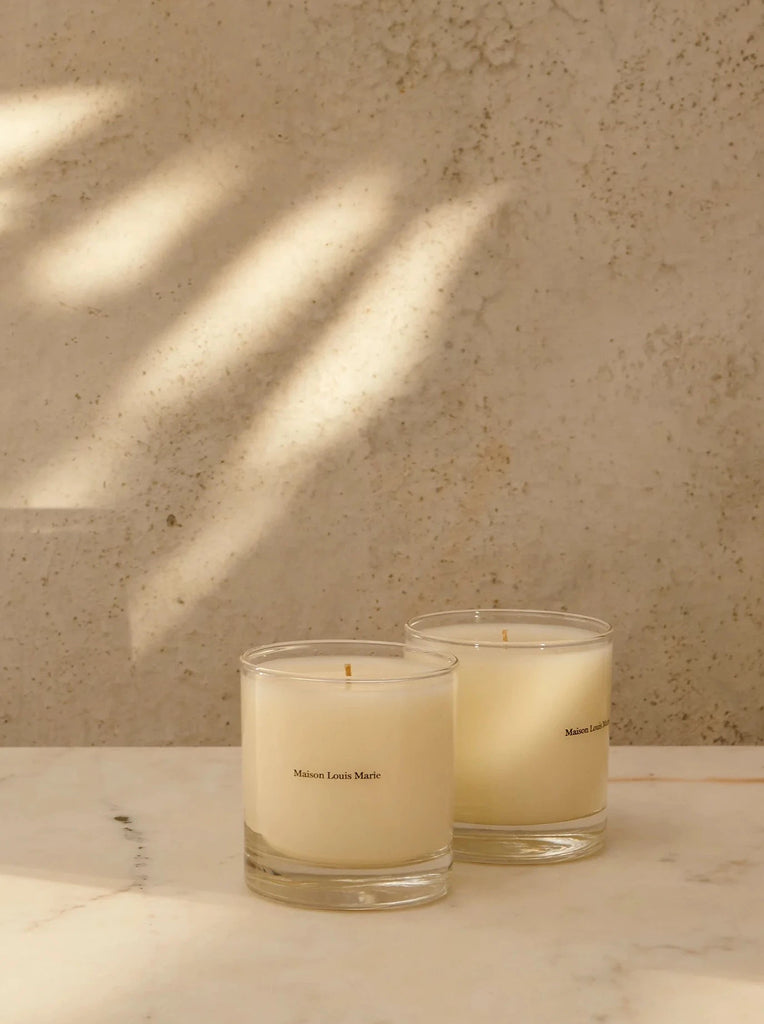 two maison louis marie candles sit in front of a tan stone background with sunlight dappling the wall.