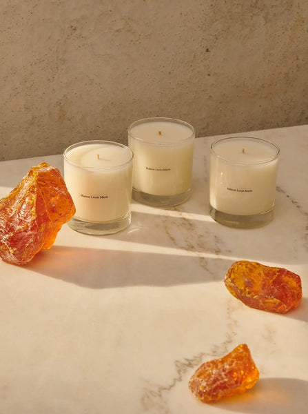 three candles from Maison Louis Marie sit on a marble table next to three pieces of amber rock.