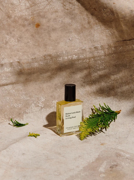a bottle of Maison Louis Marie's Le Long Fond perfume oil sits on a tan canvas next to evergreens leaves.