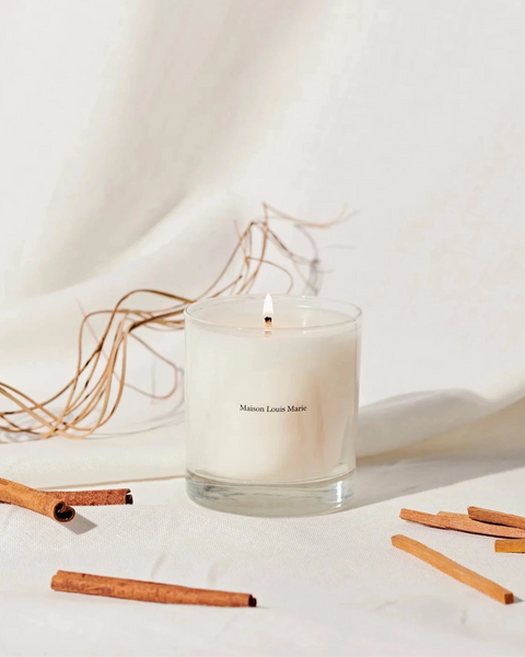 a lit candle from maison louis marie sits against a white draped background next to sandalwood and cinnamon sticks.