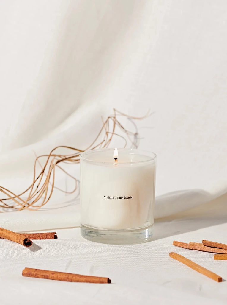 a lit candle from maison louis marie sits against a white draped background next to sandalwood and cinnamon sticks.