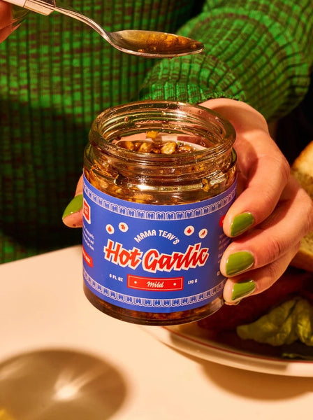 a person in a green shirt with green fingernails is holding a jar of mama teav's mild hot garlic and spoon filled with some of the garlic