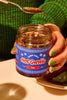 a person in a green shirt with green fingernails is holding a jar of mama teav's mild hot garlic and spoon filled with some of the garlic