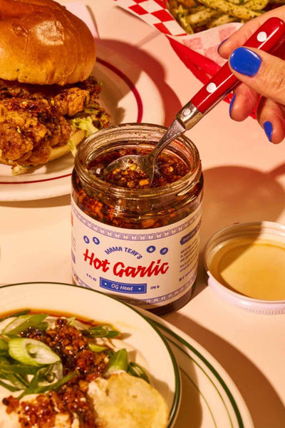 a hand with blue nail polish scooping out of a jar of mama teav's og heat hot garlic sitting among plates of food