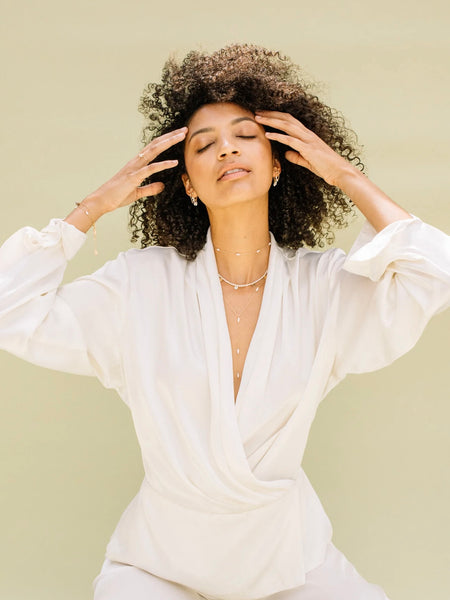 a y-necklace by Marida jewelry with freshwater seed pearls is being worn by a woman in a white jumpsuit while she sits in the sun 