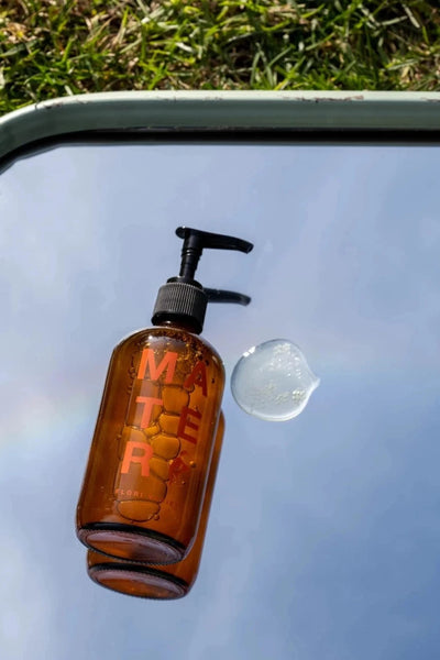 a glass bottle of flori liquid soap from mater lays next to some spilled soap on top of a mirror that reflects the blue sky